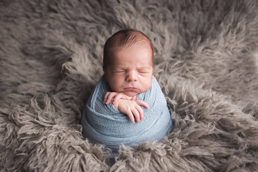 Sleeping newborn wrapped in blue swaddle on grey fur backdrop in professional studio setting.