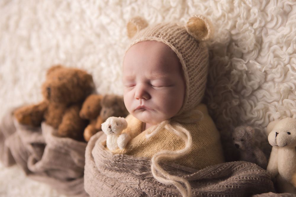 Sleeping newborn baby wearing knit bear hat and wrapped in blanket with stuffed animals.