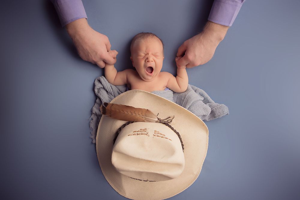 Creative newborn photo with cowboy hat and supporting hands against blue background.