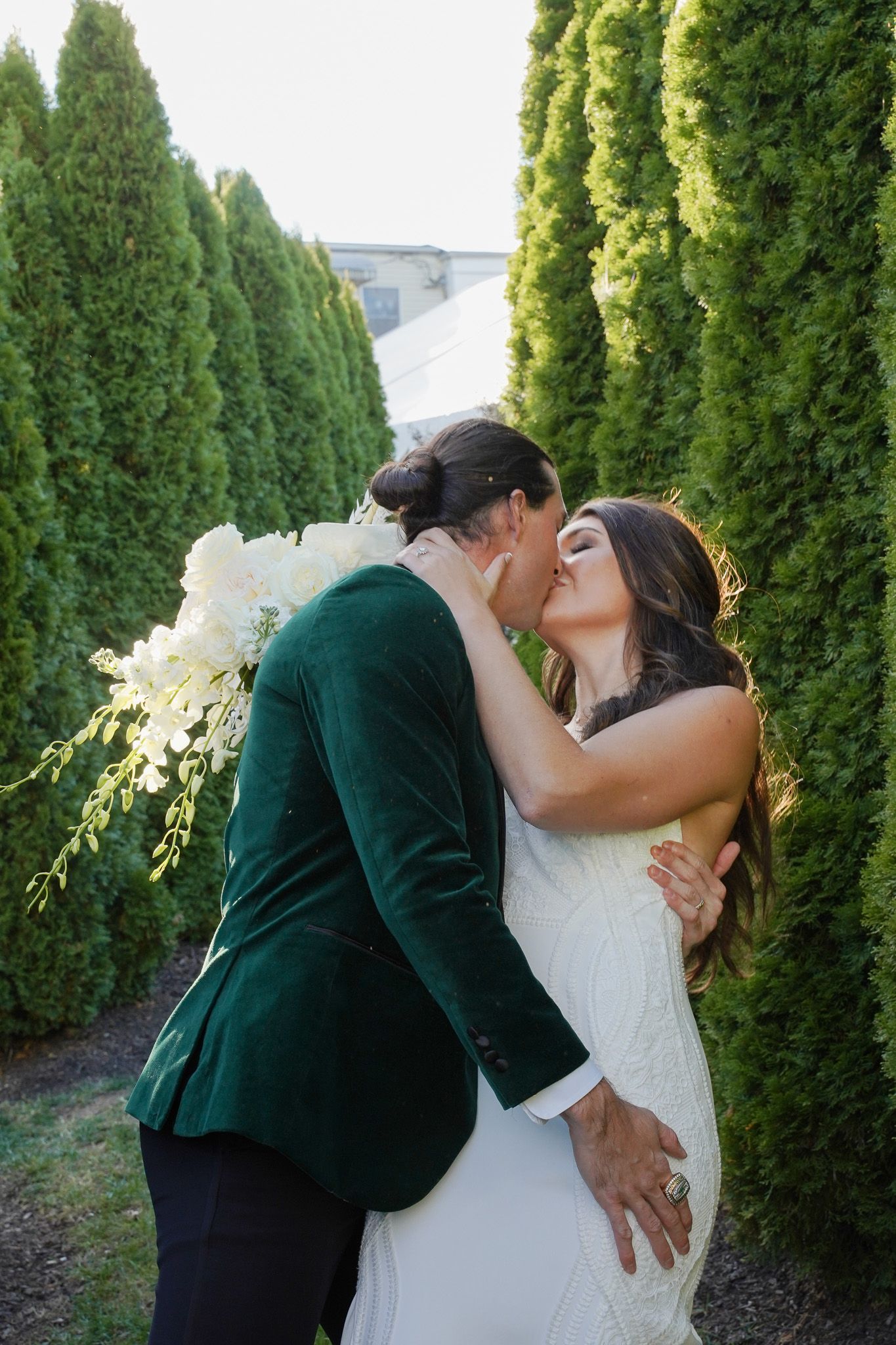 A couple shares a romantic kiss in front of tall green cypress trees on their wedding day.