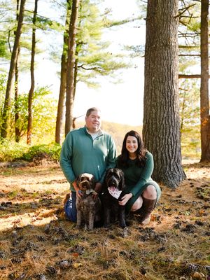 A couple in matching green sweaters poses with their two brown dogs in a sunlit autumn forest setting.