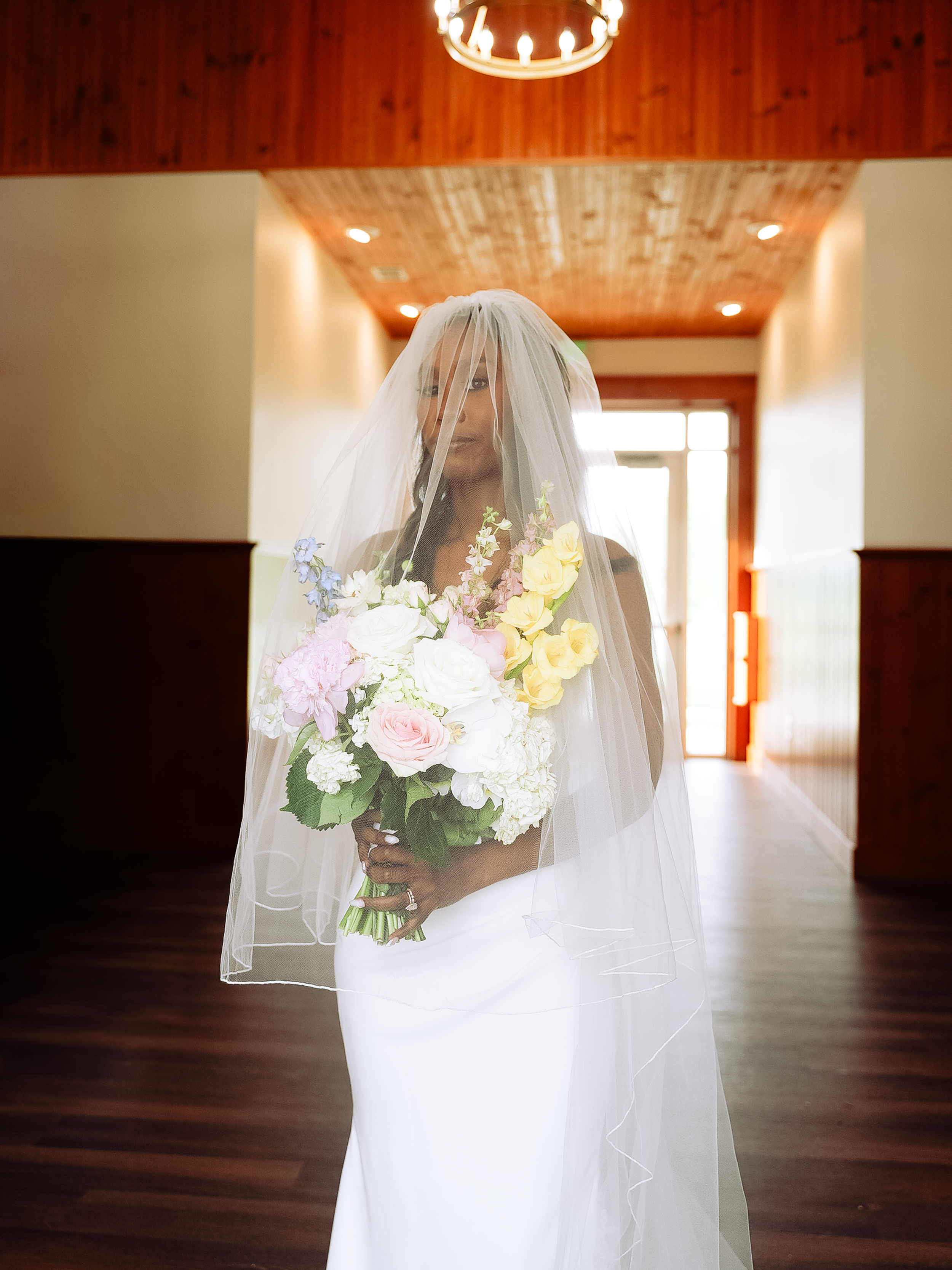 A bride in a white gown and veil holds a colorful spring bouquet in an elegant wood-paneled hallway.