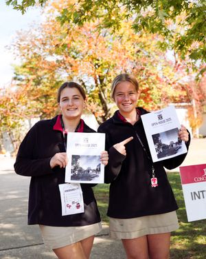 Students in matching uniforms hold up photos while standing under autumn trees with colorful foliage.