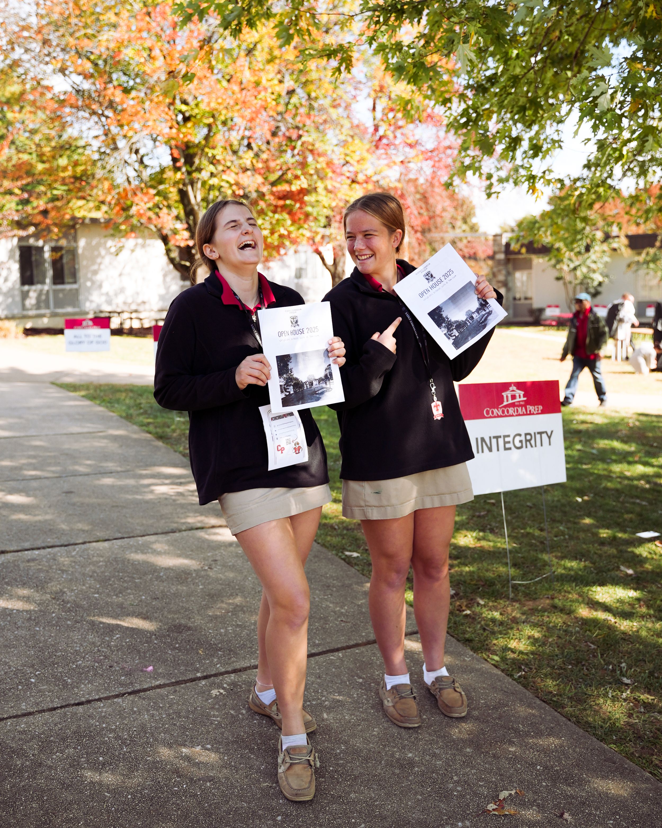 People holding signs and walking on a sidewalk path during autumn with colorful fall foliage in the background.
