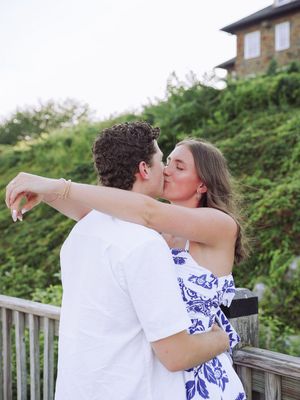 A couple in matching white and blue outfits share a romantic kiss on a wooden deck with greenery behind them.