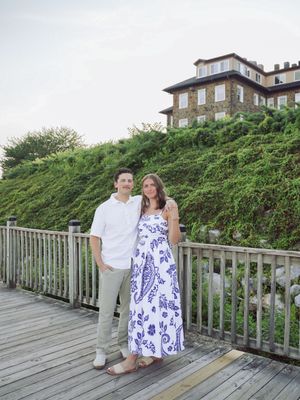 A couple stands on a wooden boardwalk overlooking the beach with green foliage and buildings in the background.