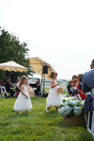Two flower girls in white dresses dance on grass at an outdoor wedding with blue hydrangeas and guests watching.