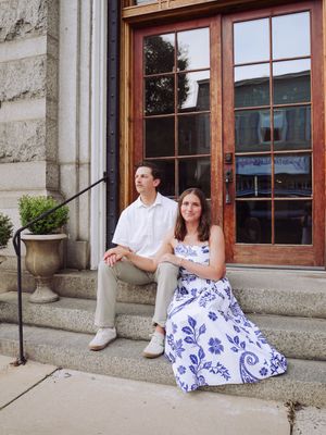 A couple sits on stone steps in front of a building with wood-framed windows wearing white and blue outfits.