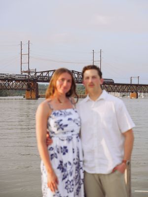 A couple stands by a waterfront with a bridge visible in the background on a cloudy day.