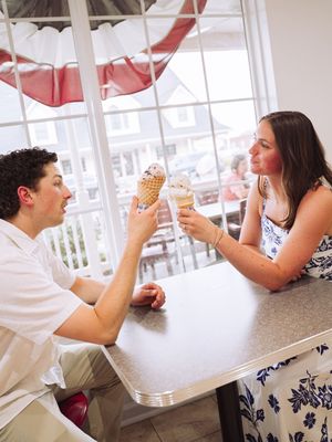 Two people share a playful moment laughing together while eating ice cream cones at an outdoor patio table.