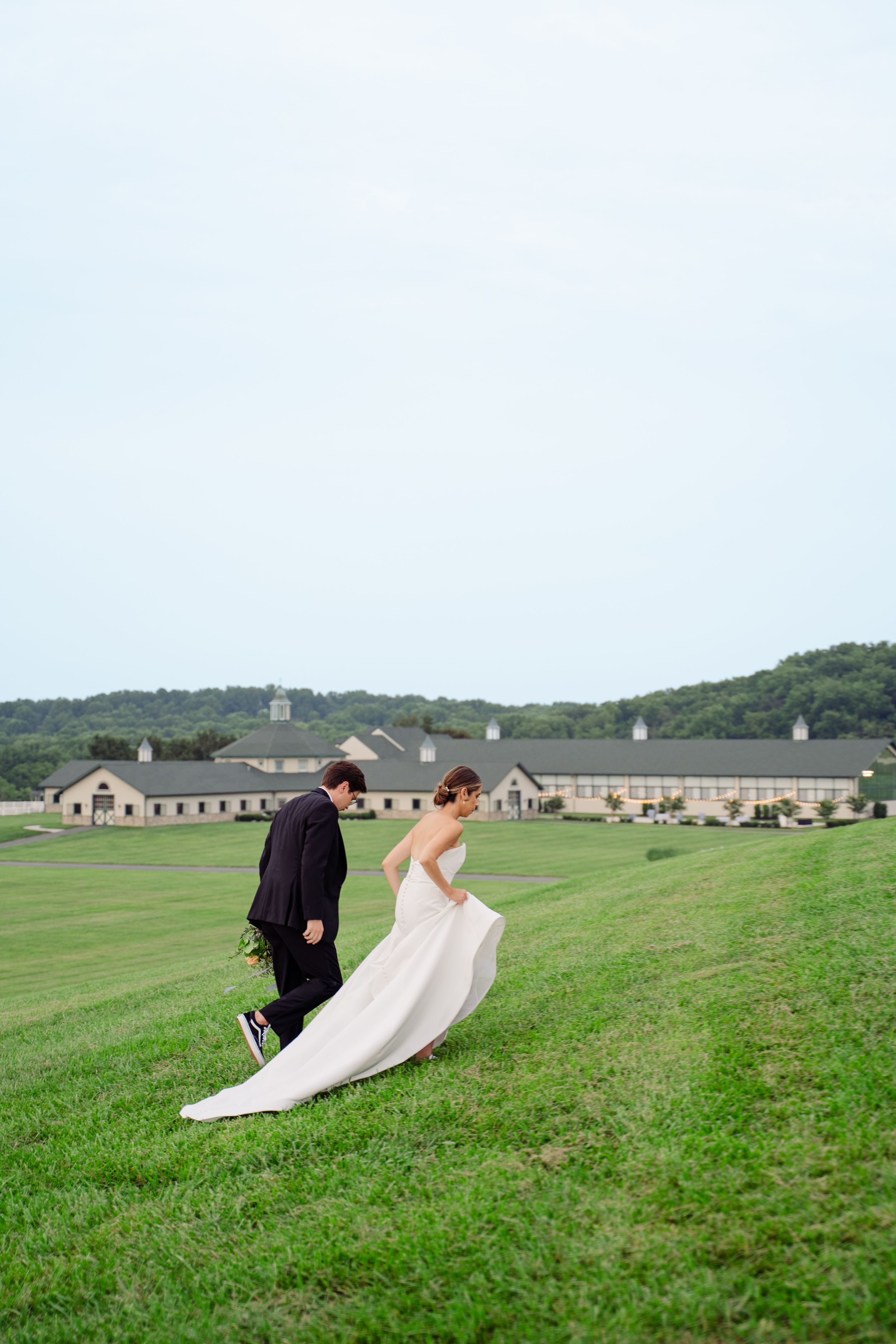 Bride and groom walking on lush green hillside, wedding dress flowing in wind, rustic venue in background.