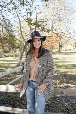 Stylish fall outfit featuring a plaid blazer, jeans and wide-brim hat against autumn foliage.