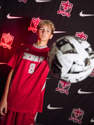A soccer player in a red Concordia Bay jersey poses against a dark backdrop with illuminated team logos and a soccer ball.