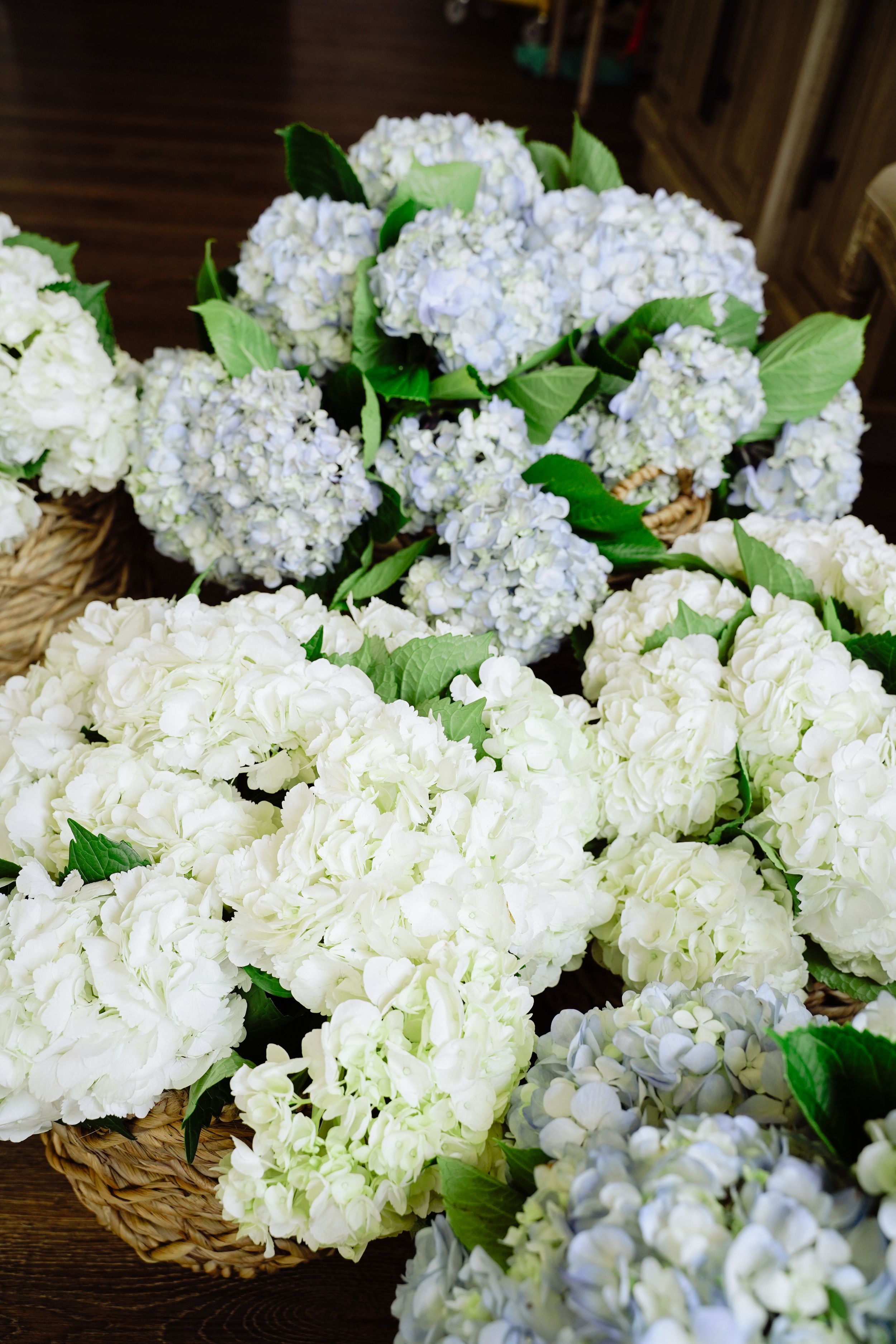 Wicker baskets filled with white and light blue hydrangea bouquets with green leaves on a wooden surface.