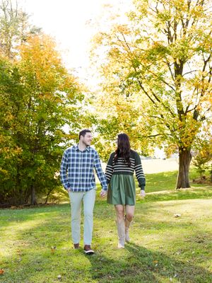 A couple walks hand in hand through a scenic autumn park with golden leaves and sunlit trees in the background.