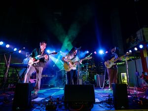 A band performs on stage with colorful blue and green lighting effects illuminating their live performance setup.