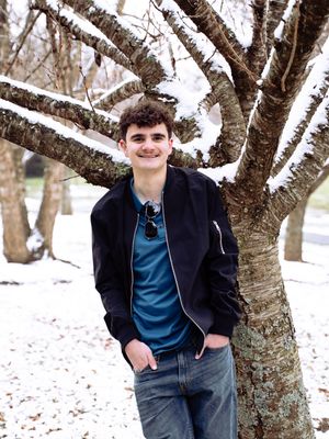 Person in dark jacket and blue shirt poses casually against snow-covered trees in winter landscape.