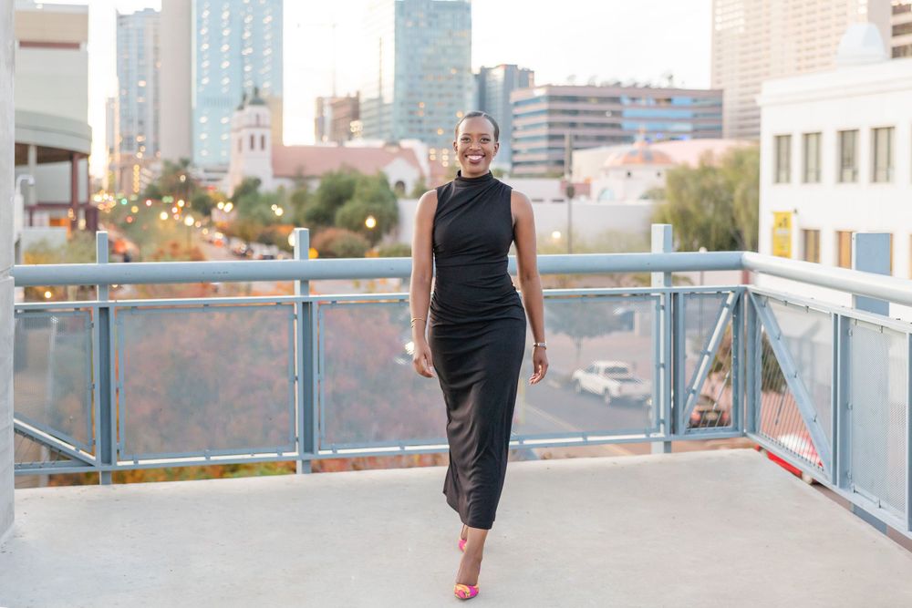 Woman in black sleeveless dress poses confidently on city rooftop terrace.