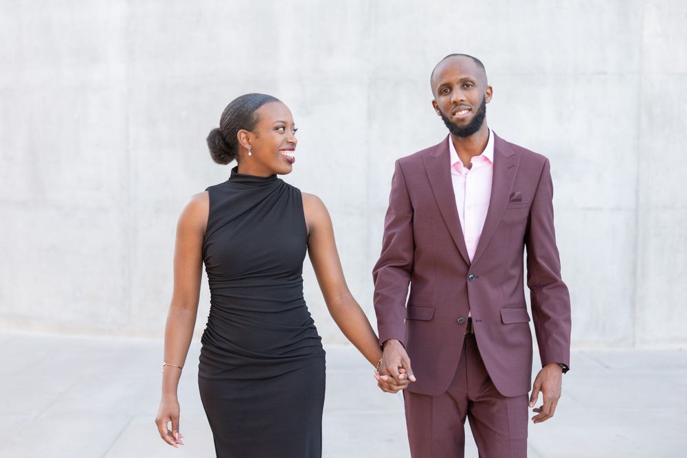 Couple in formal attire poses against white wall, wearing black dress and burgundy suit.