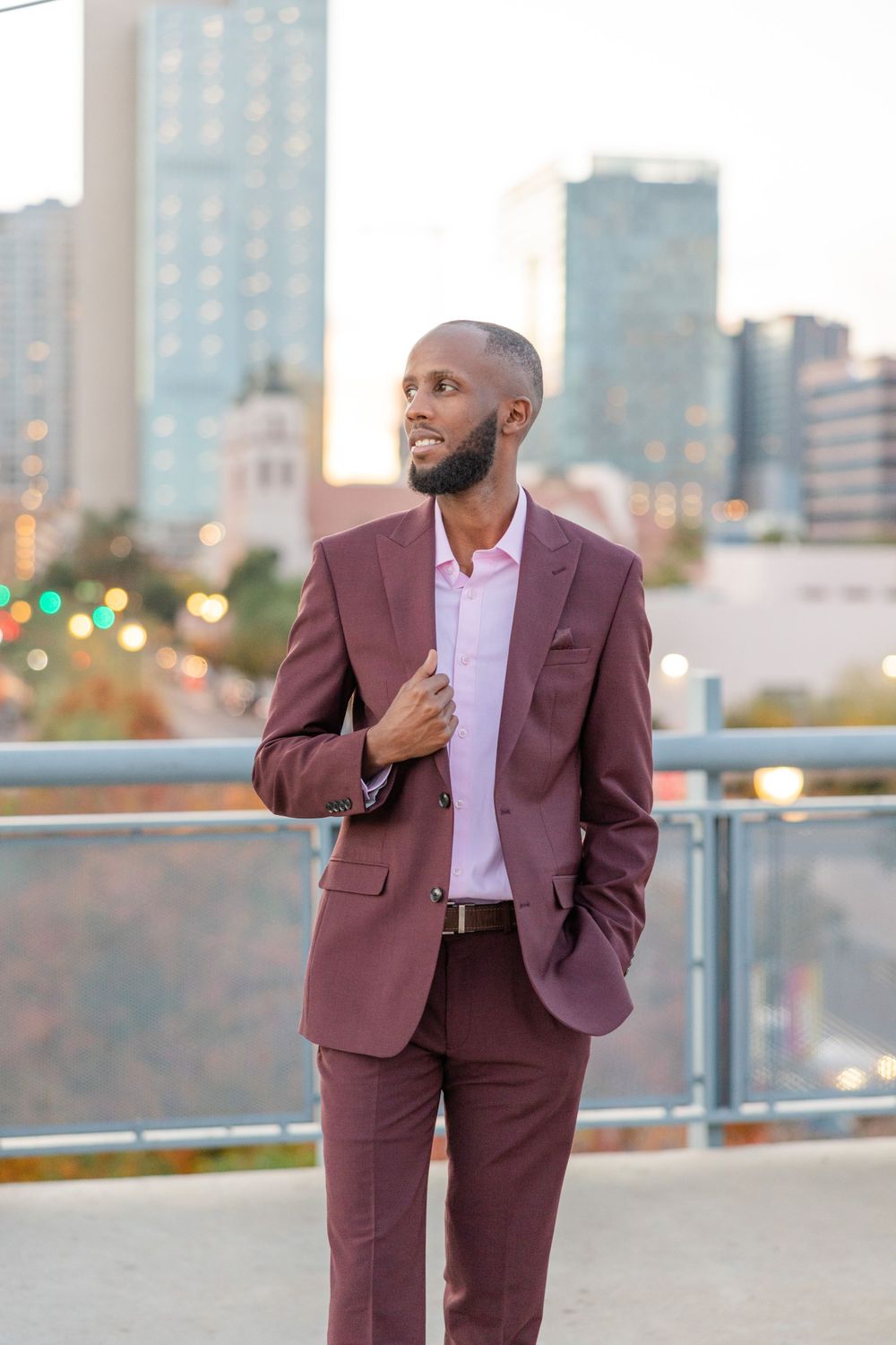 A stylish man in a burgundy suit poses on an urban rooftop at sunset.