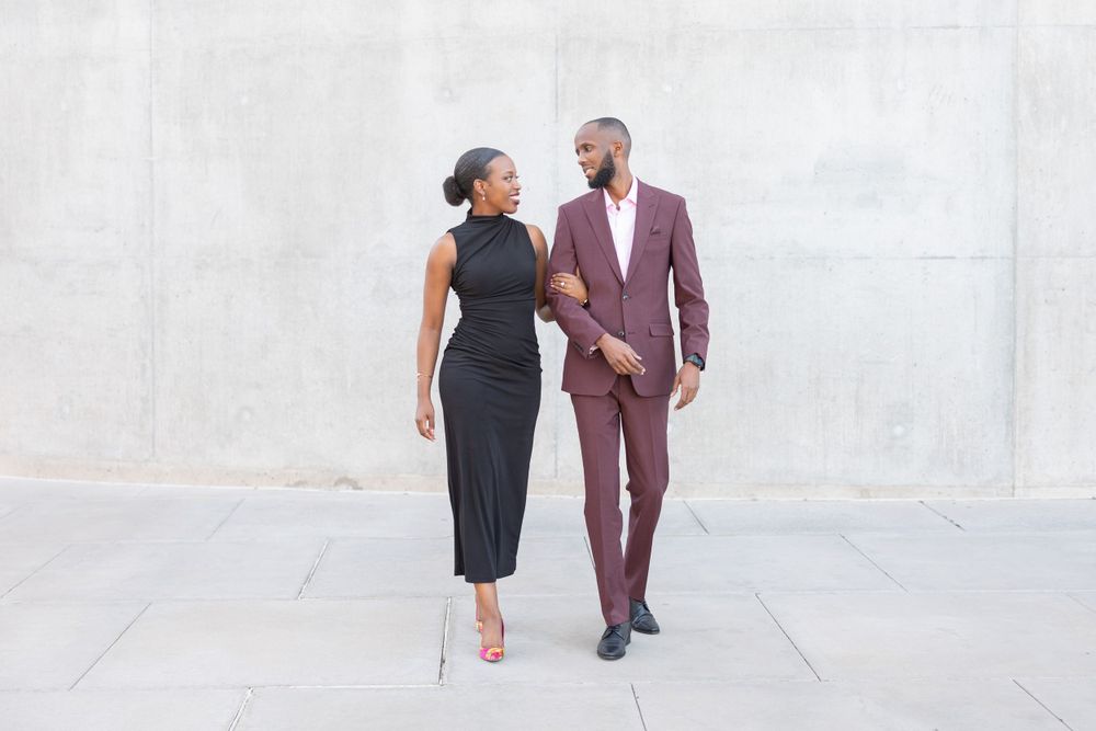 A couple in formal attire walks together, with her in a black dress and him in a burgundy suit.