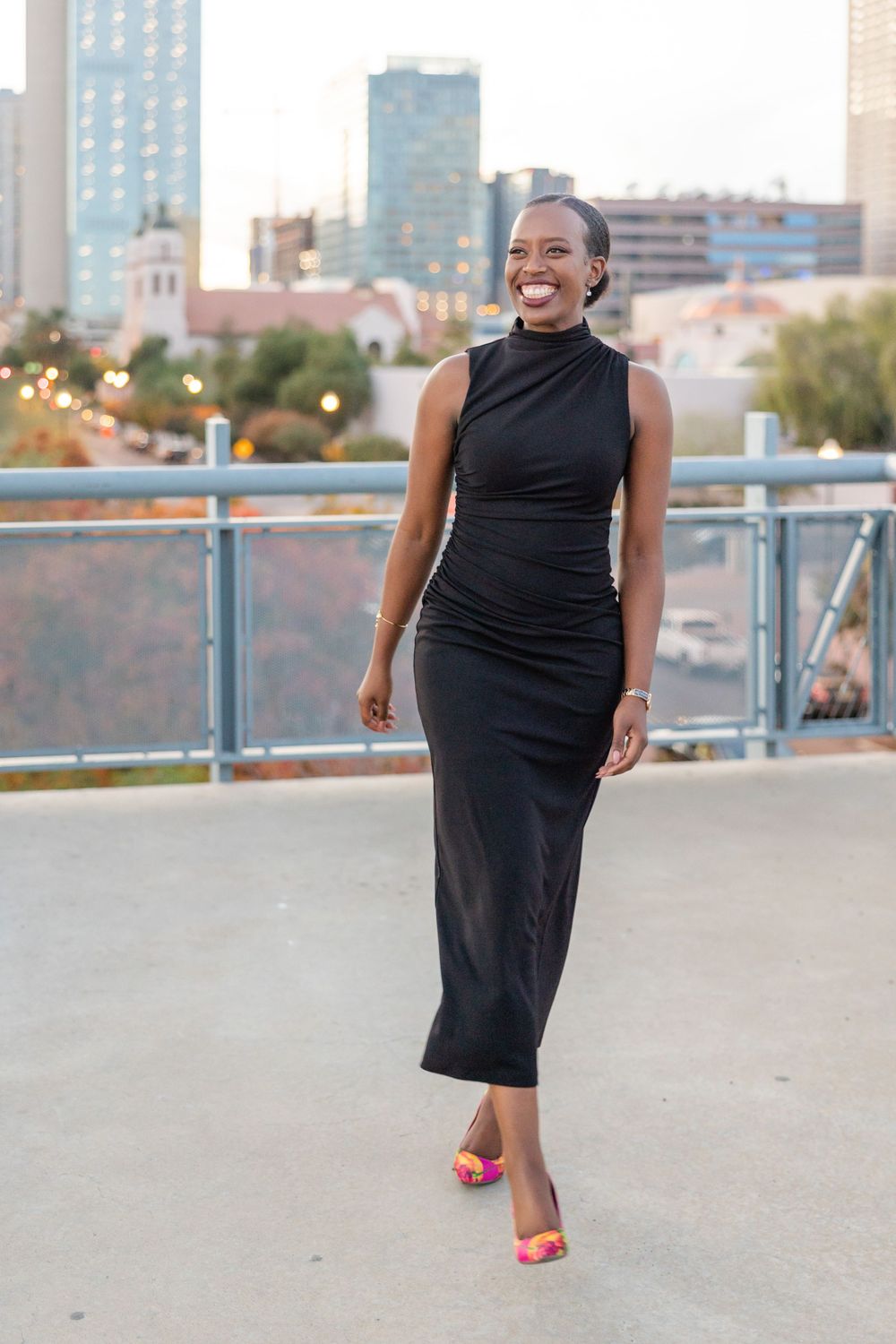 Elegant woman in black sleeveless dress and red shoes stands in front of an urban cityscape at dusk.