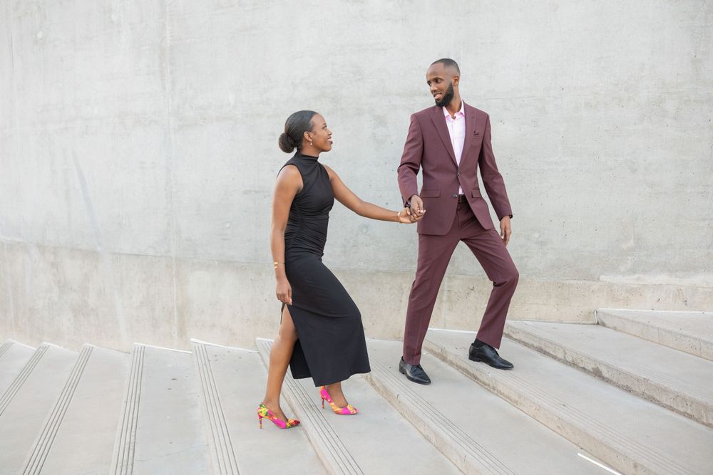 A couple playfully poses on concrete steps, with her in a black dress and him in a burgundy suit.