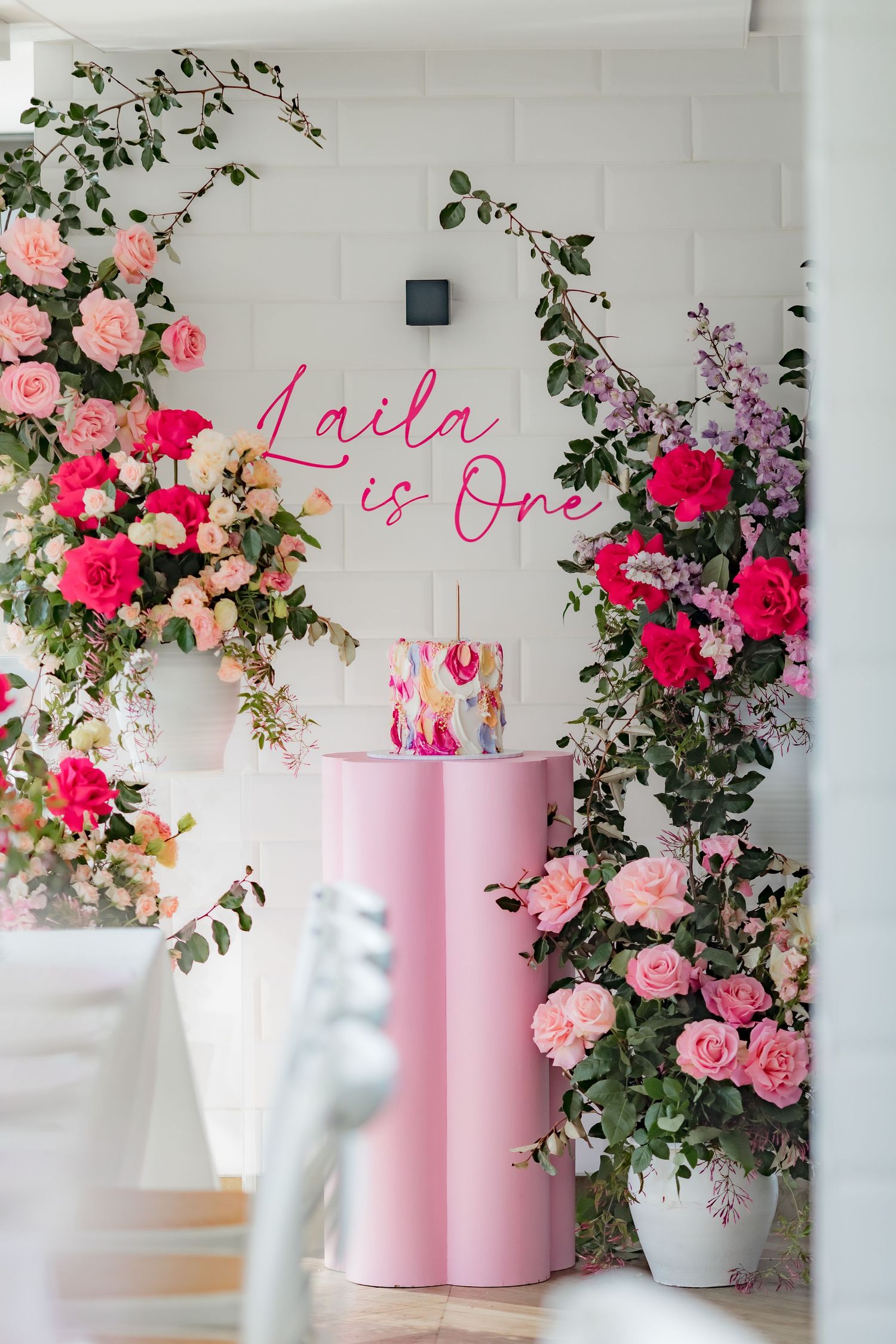 Pink neon birthday sign surrounded by lush floral arrangements against a white brick wall.