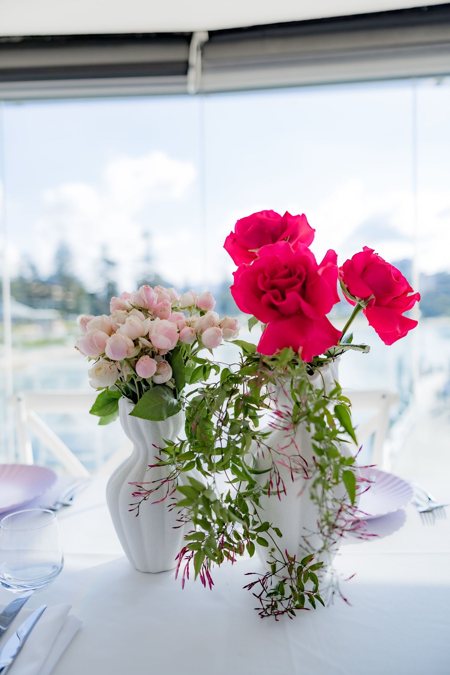 Vibrant red and pink roses in a white vase with waterfront view in background.