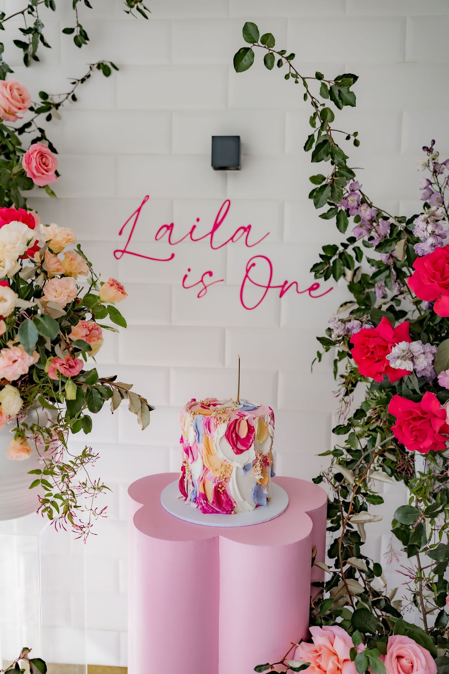 Pink neon 'Laila is One' sign above watercolor cake surrounded by roses and greenery.