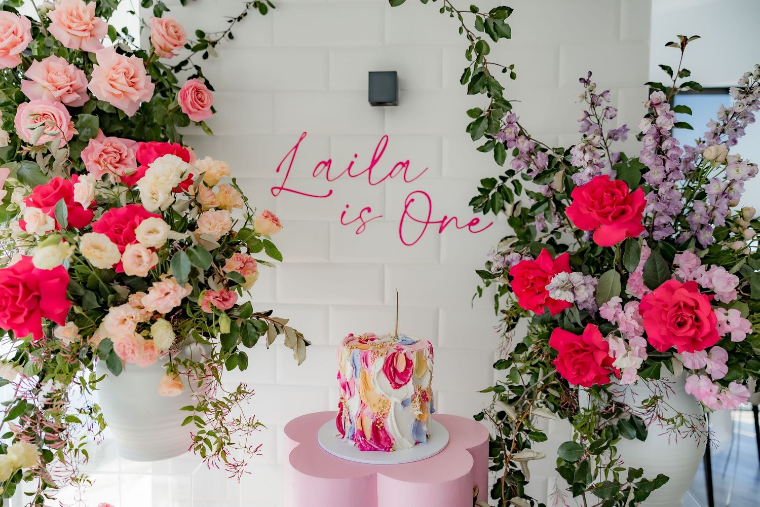First birthday display featuring watercolor cake on pink stand with floral arrangements and wall signage.