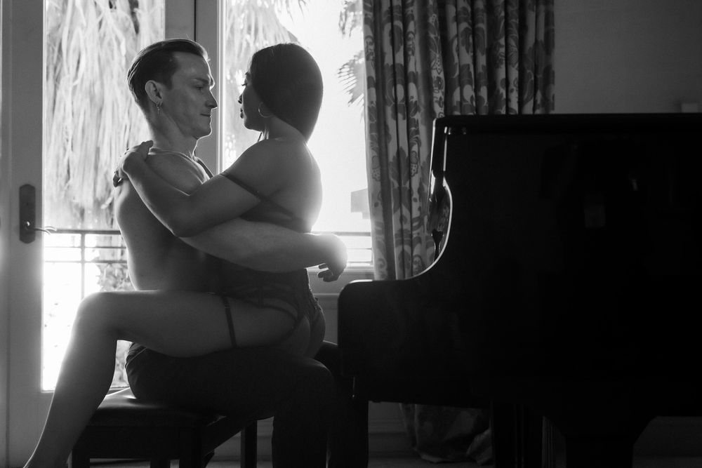Black and white silhouette of a couple embracing intimately beside a grand piano near a sunlit window.