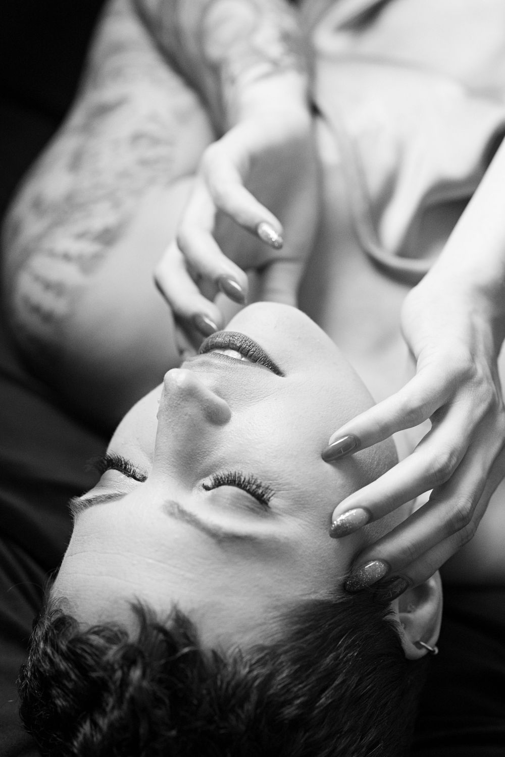 Black and white intimate portrait of a woman lying back with eyes closed while a hand gently touches her face.