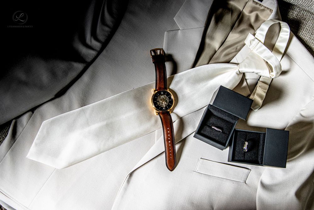 Wedding accessories including rings boxes and brown leather watch on white suit.
