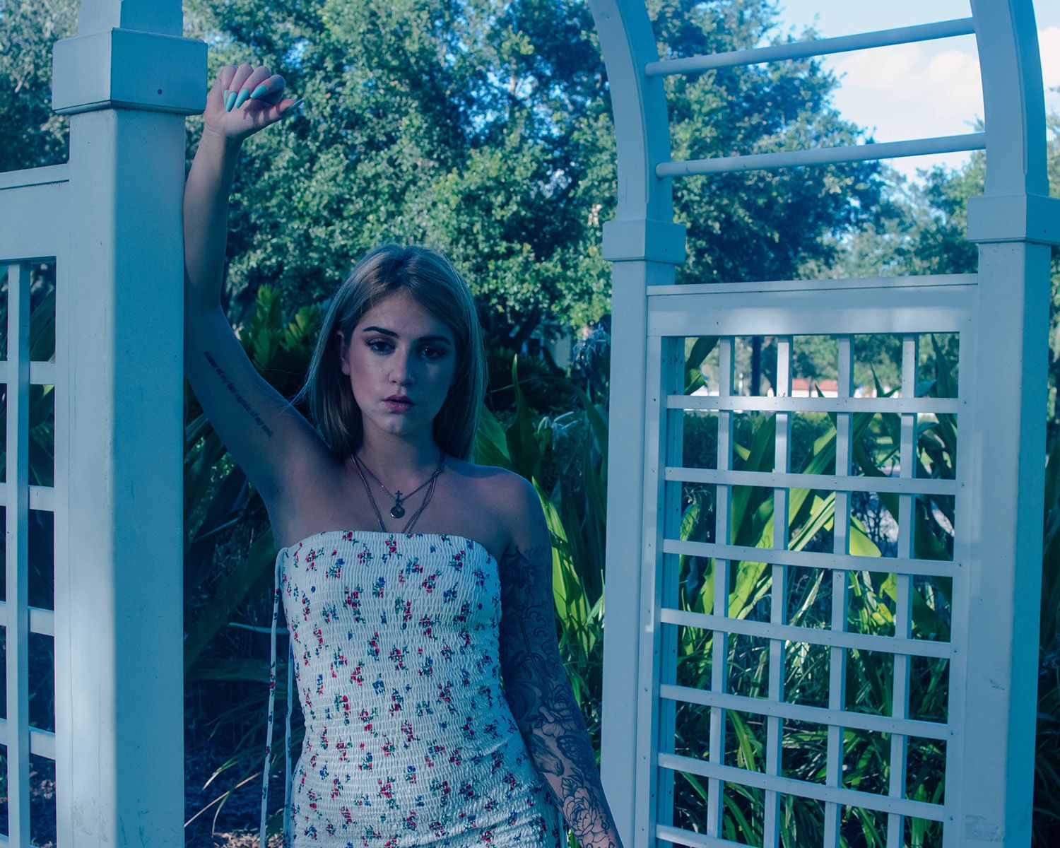 Person in floral dress poses elegantly by garden trellis in evening light.