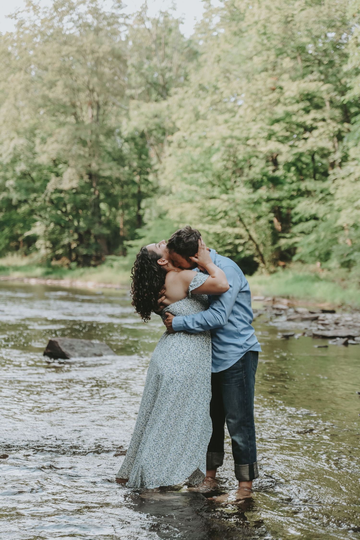 A romantic couple dances in a shallow creek surrounded by lush greenery on a sunny day.