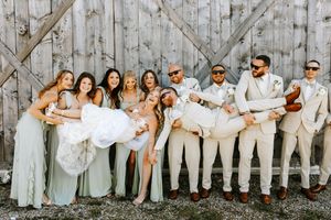 Wedding party members in light colored attire pose casually against a rustic wooden fence outdoors.