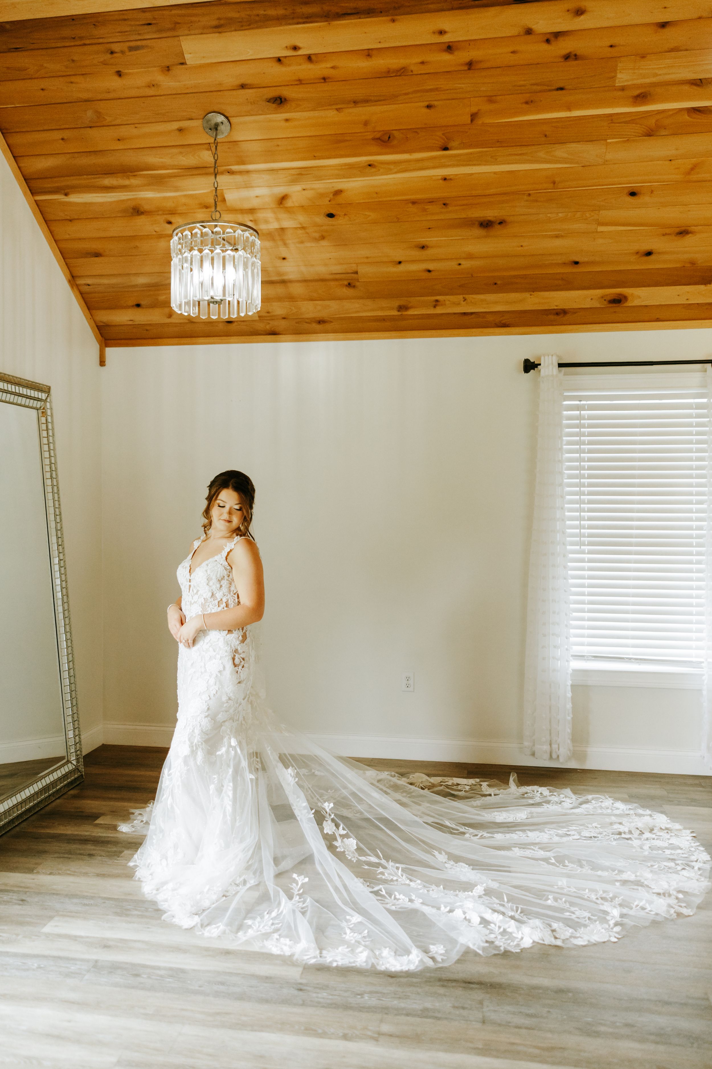 A stunning white lace wedding gown with a flowing cathedral train set against a room with wooden ceiling and crystal chandelier.