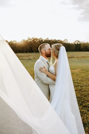 A romantic wedding couple embraces in a sunlit field as the bride's long white veil flows dramatically in the breeze.
