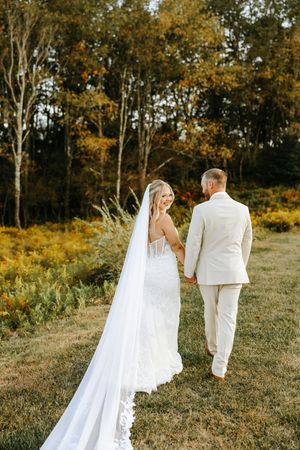 A bride and groom walk hand in hand through a sunlit autumn field with golden foliage in the background.