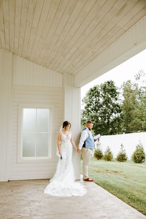 A wedding couple stands outside a white chapel building with greenery in the background.