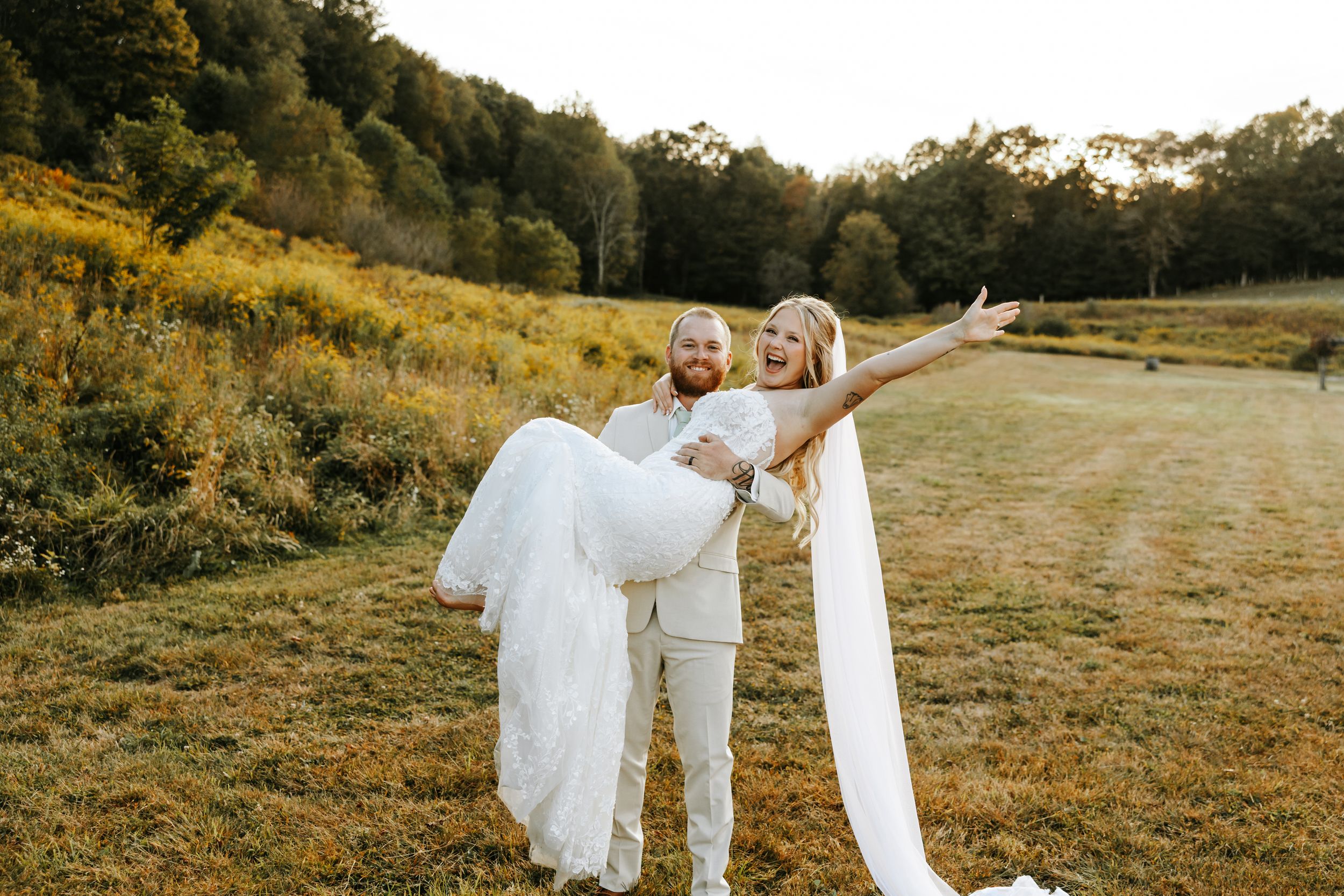 A couple in wedding attire dances together in a golden field at sunset during their outdoor celebration.