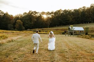 Couple walks hand in hand through a golden sunlit meadow at sunset with a rustic barn in the distance.