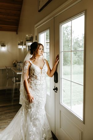 A bride in a white lace wedding gown with floral appliques poses near a sunlit window in a rustic venue.