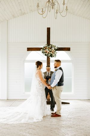 A wedding ceremony taking place in a white chapel with a wooden cross and floral arrangement.