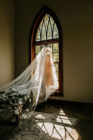 Flowing white wedding veil catches sunlight streaming through arched church window creating ethereal shadows on floor.