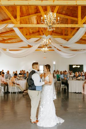 A couple shares their first dance in an elegant barn venue decorated with white drapes and crystal chandeliers.