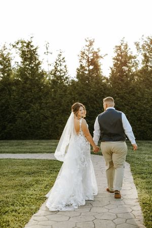 A newlywed couple walks together down a stone path surrounded by green trees at sunset.