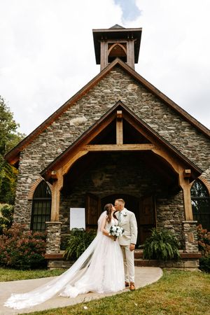 Stone chapel with wooden beams and cross sits on grassy hill during cloudy day.