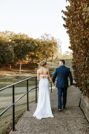 Newlyweds walking away on pathway with autumn foliage backdrop at sunset.